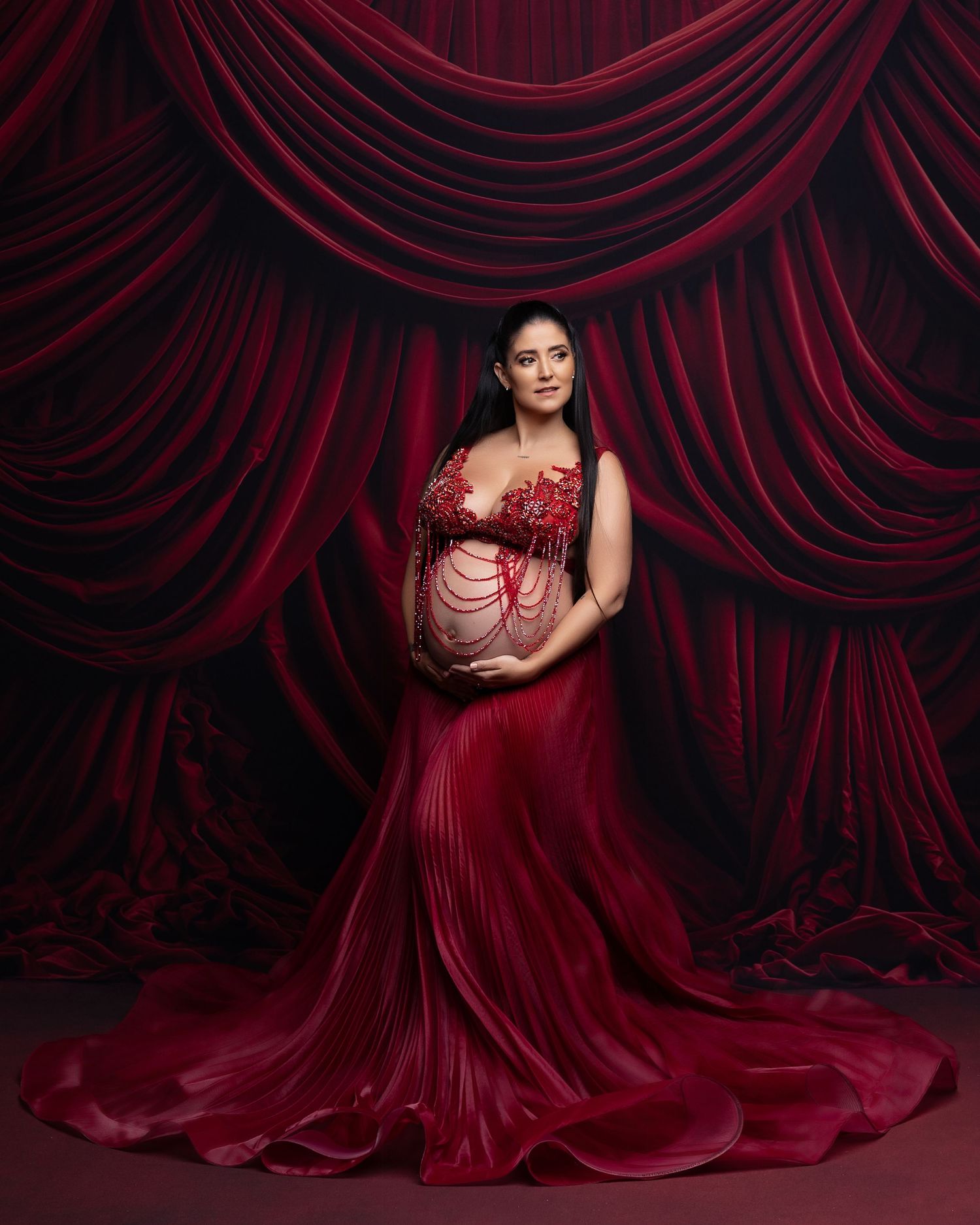 A figure in a flowing deep red gown poses dramatically against dark red velvet curtain backdrop.