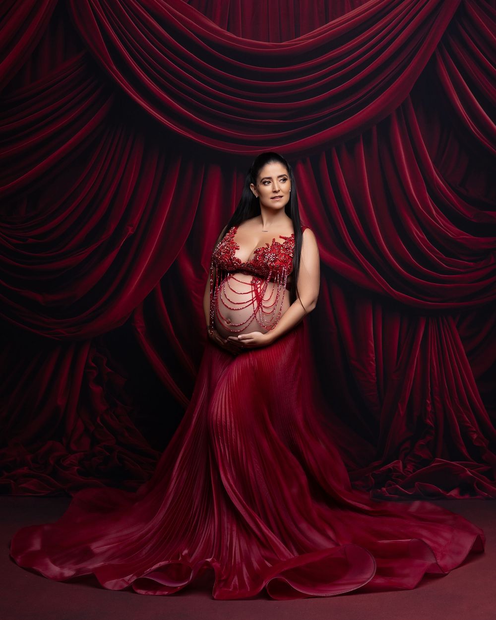 A figure in a flowing deep red gown poses dramatically against dark red velvet curtain backdrop.