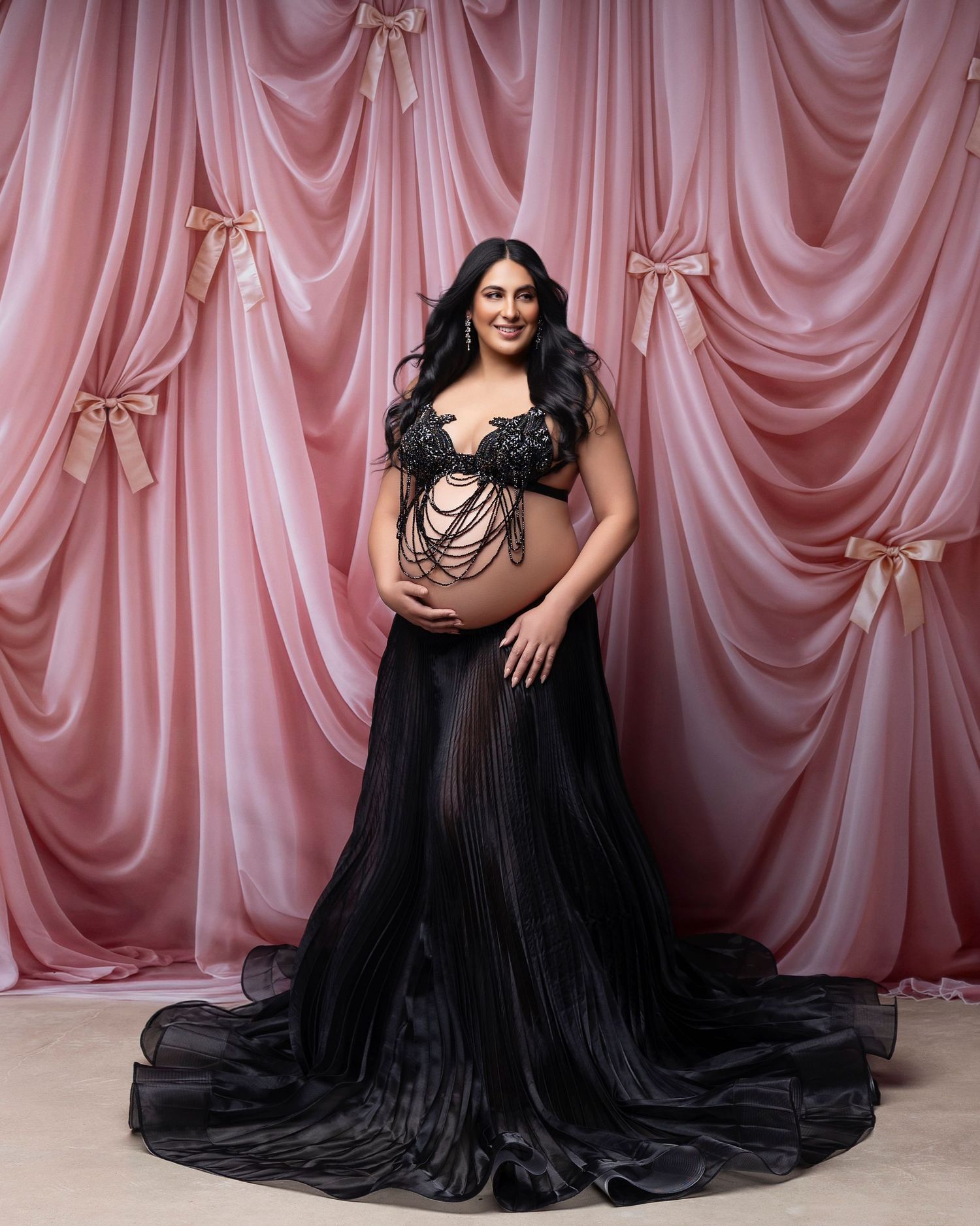 Pregnant woman in black sheer maternity gown poses against pink draped backdrop with bow accents in elegant studio photo.