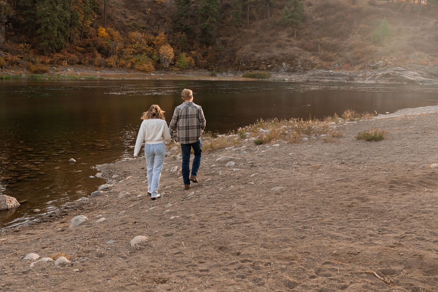 Two people walk together along a serene riverbank path surrounded by autumn foliage during golden hour.