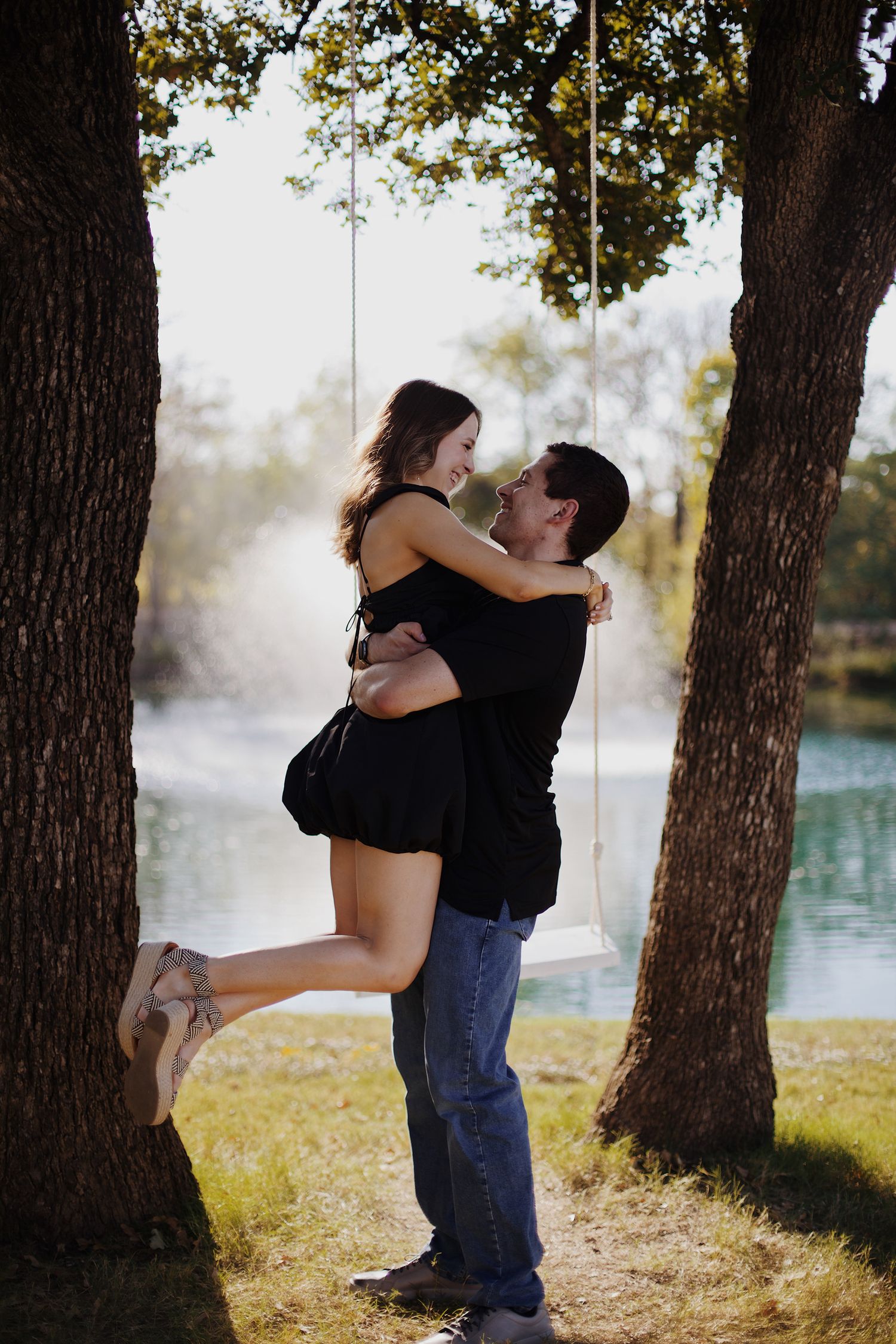 Engaged couple embracing and holding each other near a swing by the pond.