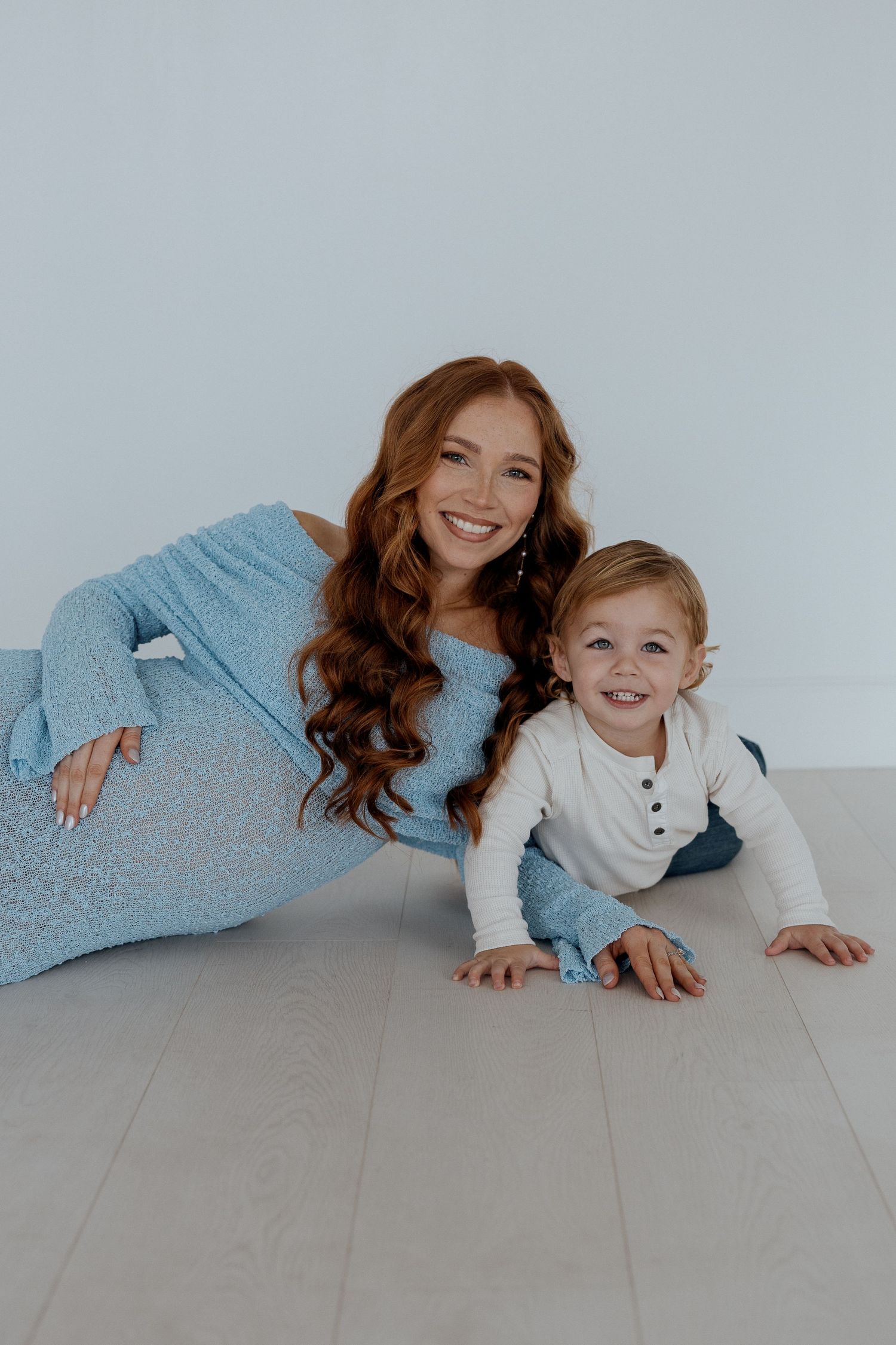 Parent and child laying on hardwood floor wearing cozy blue and white outfits while smiling at camera.