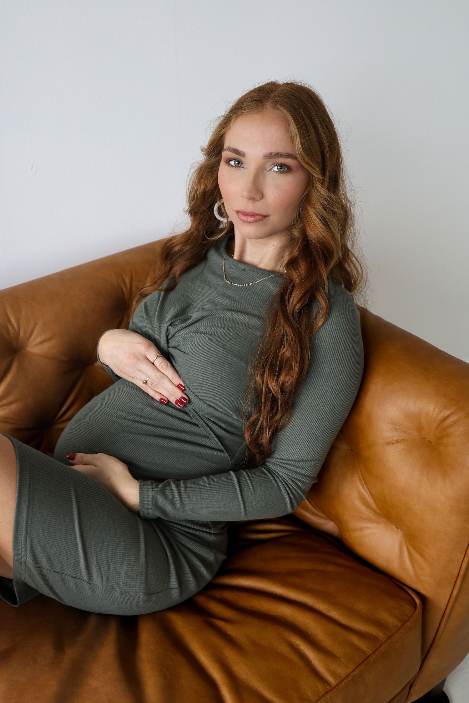 A pregnant woman in a sage green dress relaxes on a brown leather couch while posing for a portrait. Maternity, Motherhood, Studio