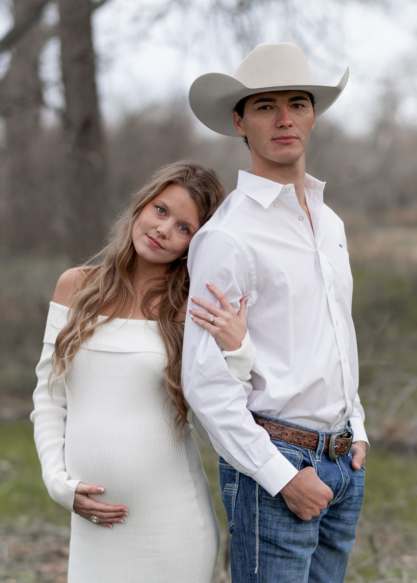 A couple in matching white outfits poses for a maternity photo outdoors against a blurred natural background.