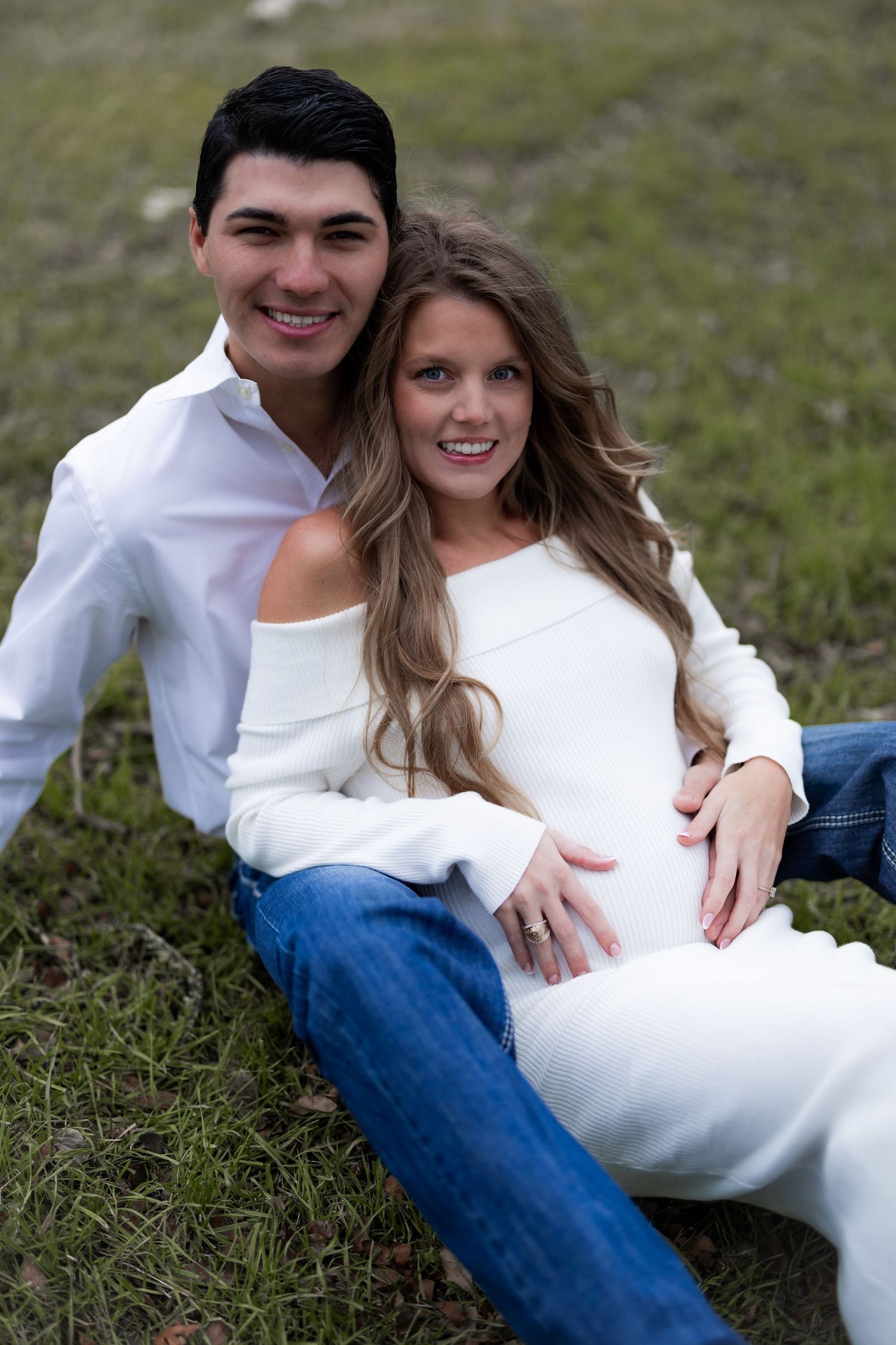 A couple in white clothing sit together on grassy ground during a romantic outdoor photoshoot.