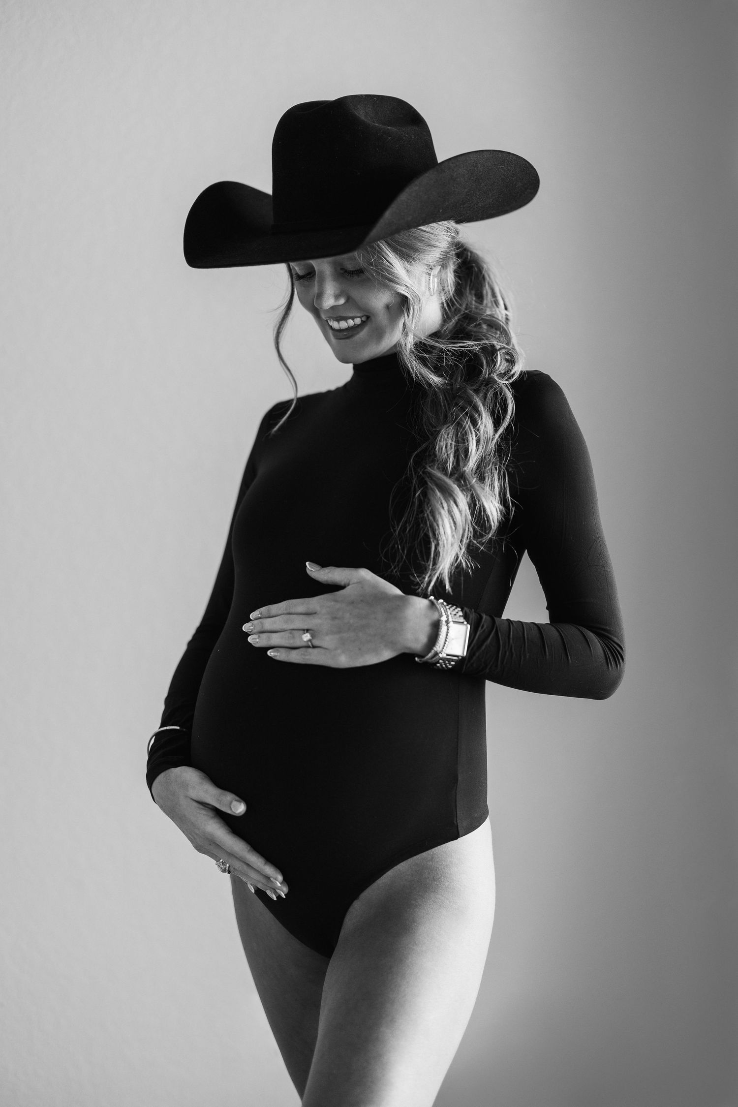 A maternity portrait in black and white showing a pregnant woman in a bodysuit and cowboy hat.