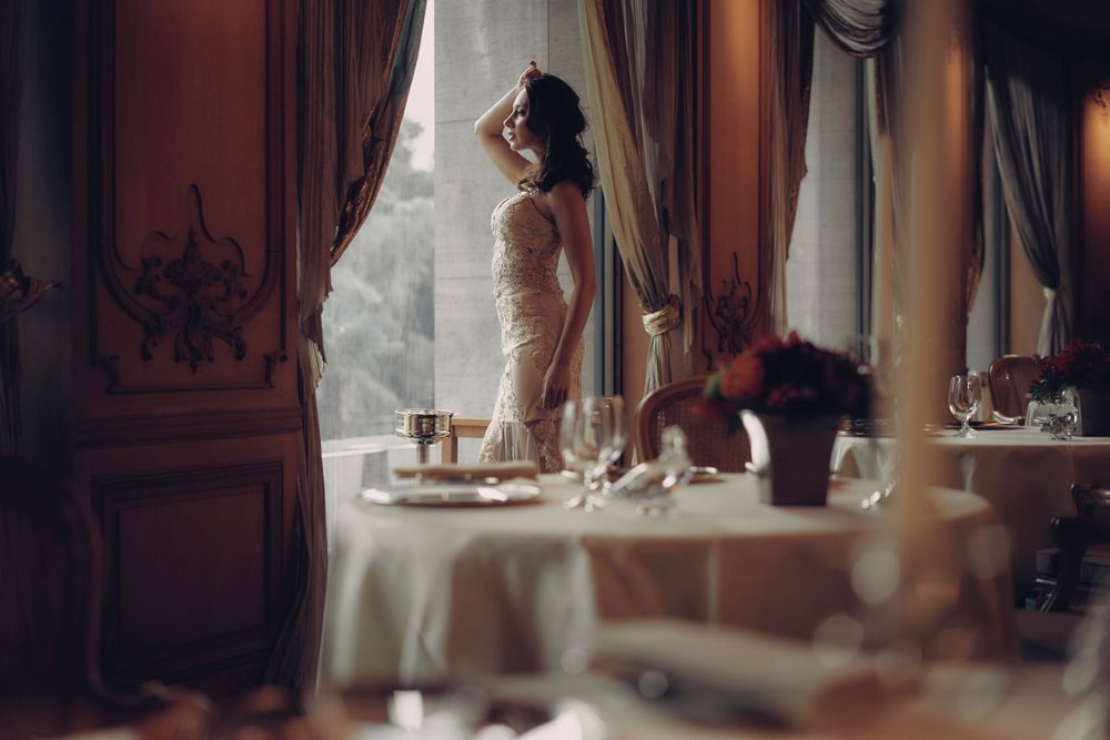 Figure in elegant white dress stands silhouetted against window in luxurious dining room with set tables. Wedding photographer
