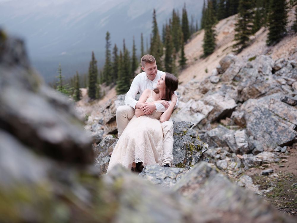 A couple in white wedding attire sits intimately among rocky mountain terrain surrounded by evergreen trees in the Canadian Rockies in Banff