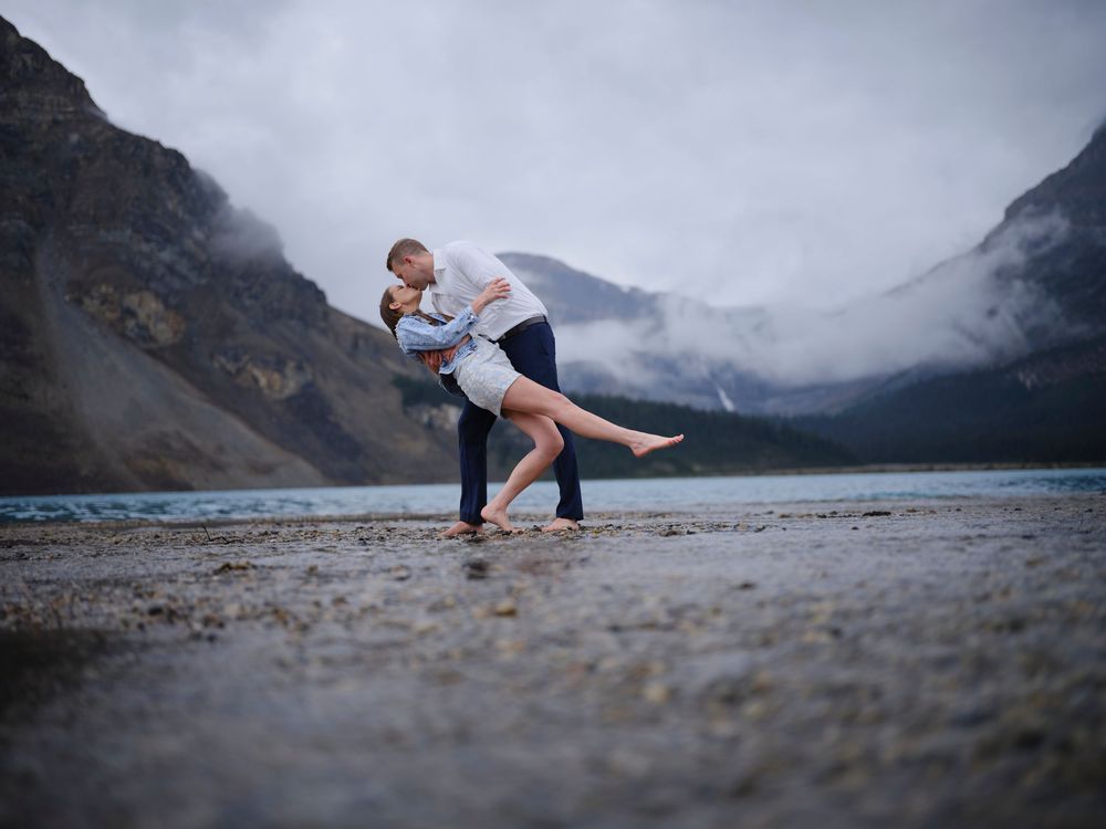 A couple dances together on a rocky beach shore with misty mountains and a lake in the dramatic background in the Canadian Rockies