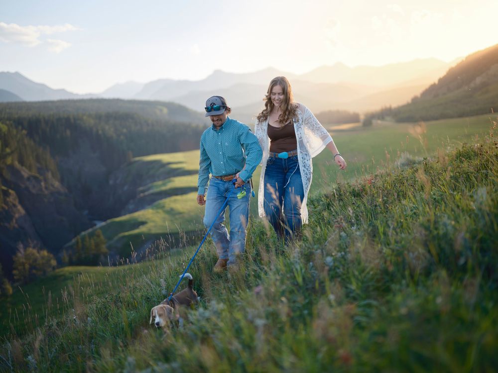 A couple walk along a grassy mountain trail at sunset with dramatic mountain landscape views in the background in the Canadian Rockies