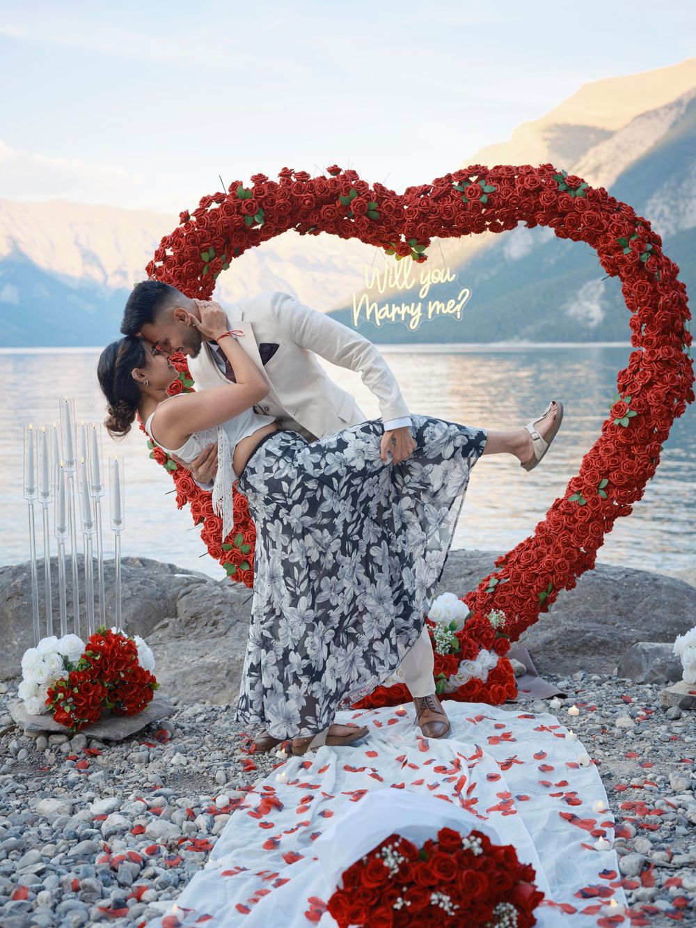 A romantic beach proposal setup with a heart-shaped arch made of red roses against a scenic mountain lake backdrop in Banff