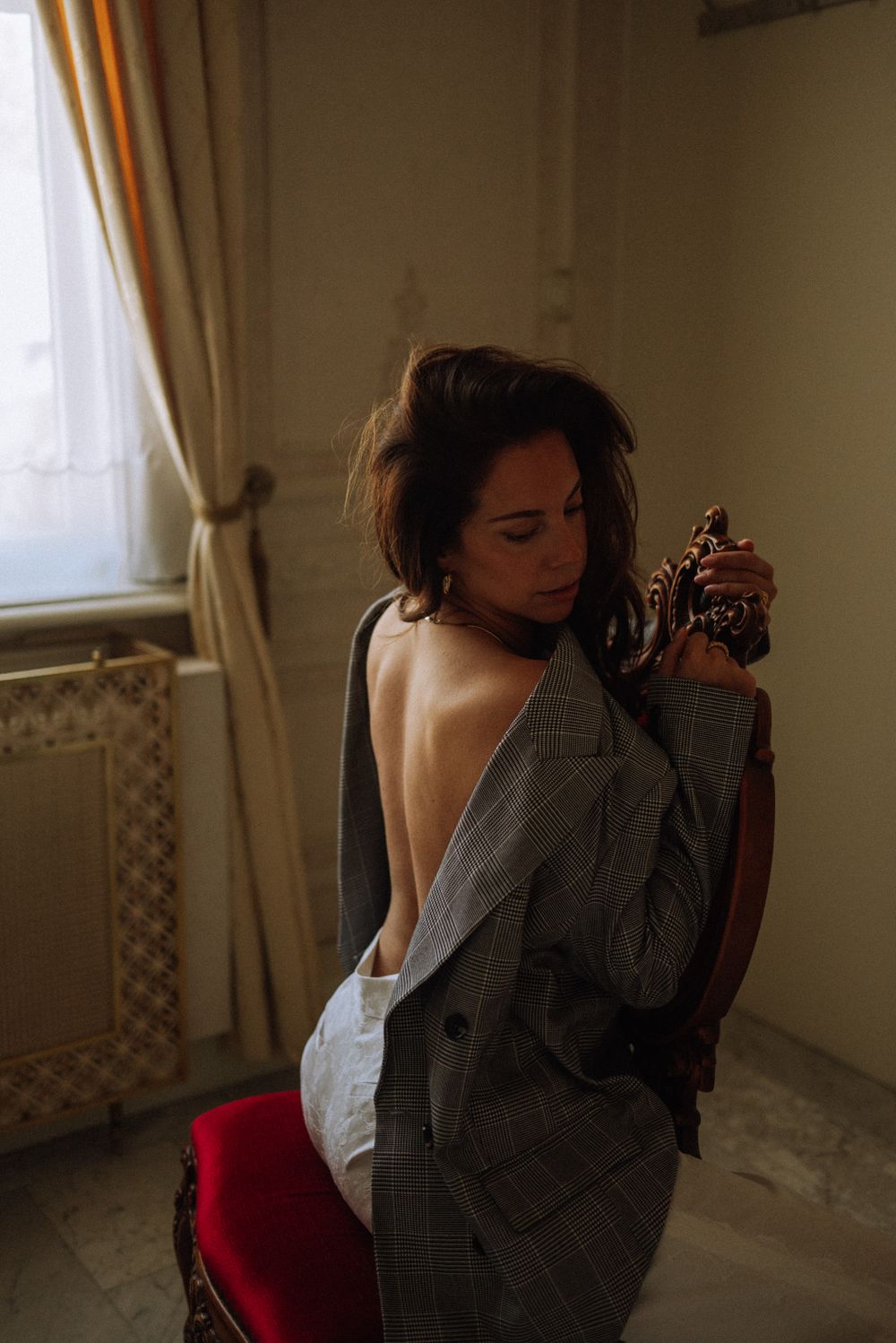 Moody boudoir portrait showing bare back in oversized blazer near window.