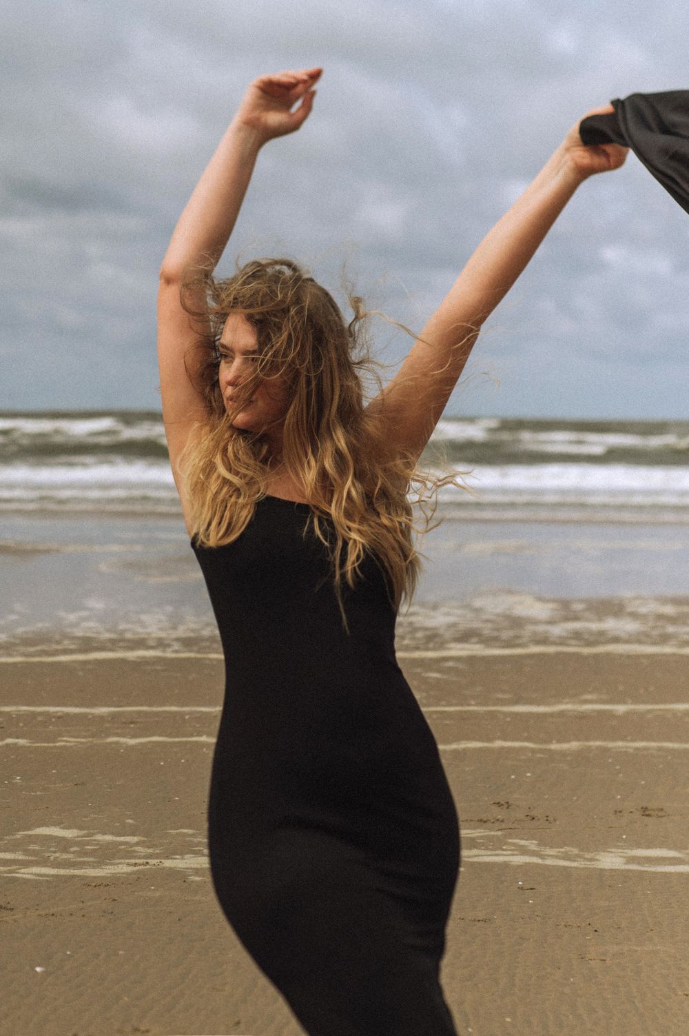 Woman in black dress dancing joyfully on a beach with arms raised.