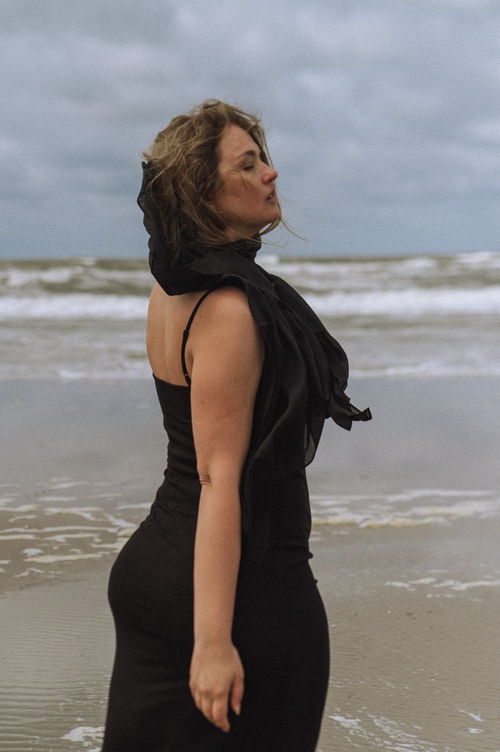 Figure in a black dress poses dramatically on a windswept beach.