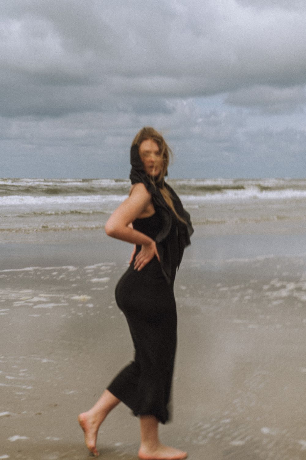 A person in a black dress dances joyfully along a cloudy beach shoreline.