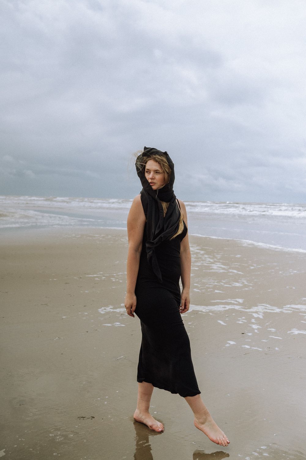 Figure in black dress walks and dances along the ocean shoreline on an overcast day.