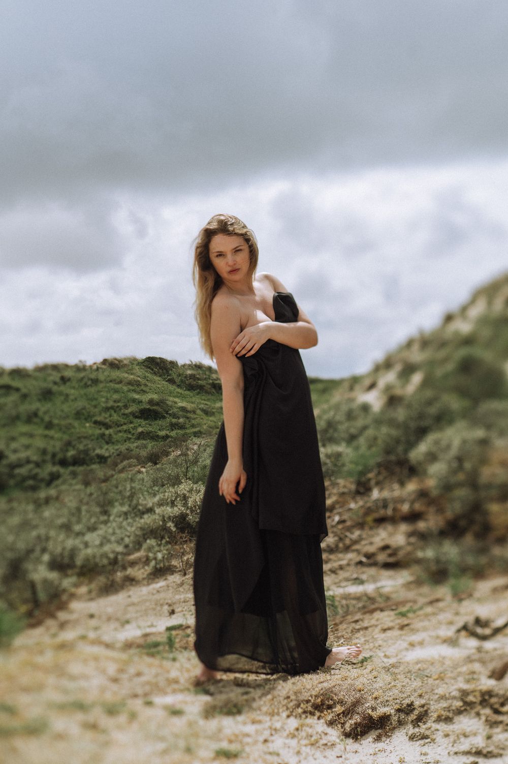 Figure in black dress poses on coastal dunes under cloudy skies.