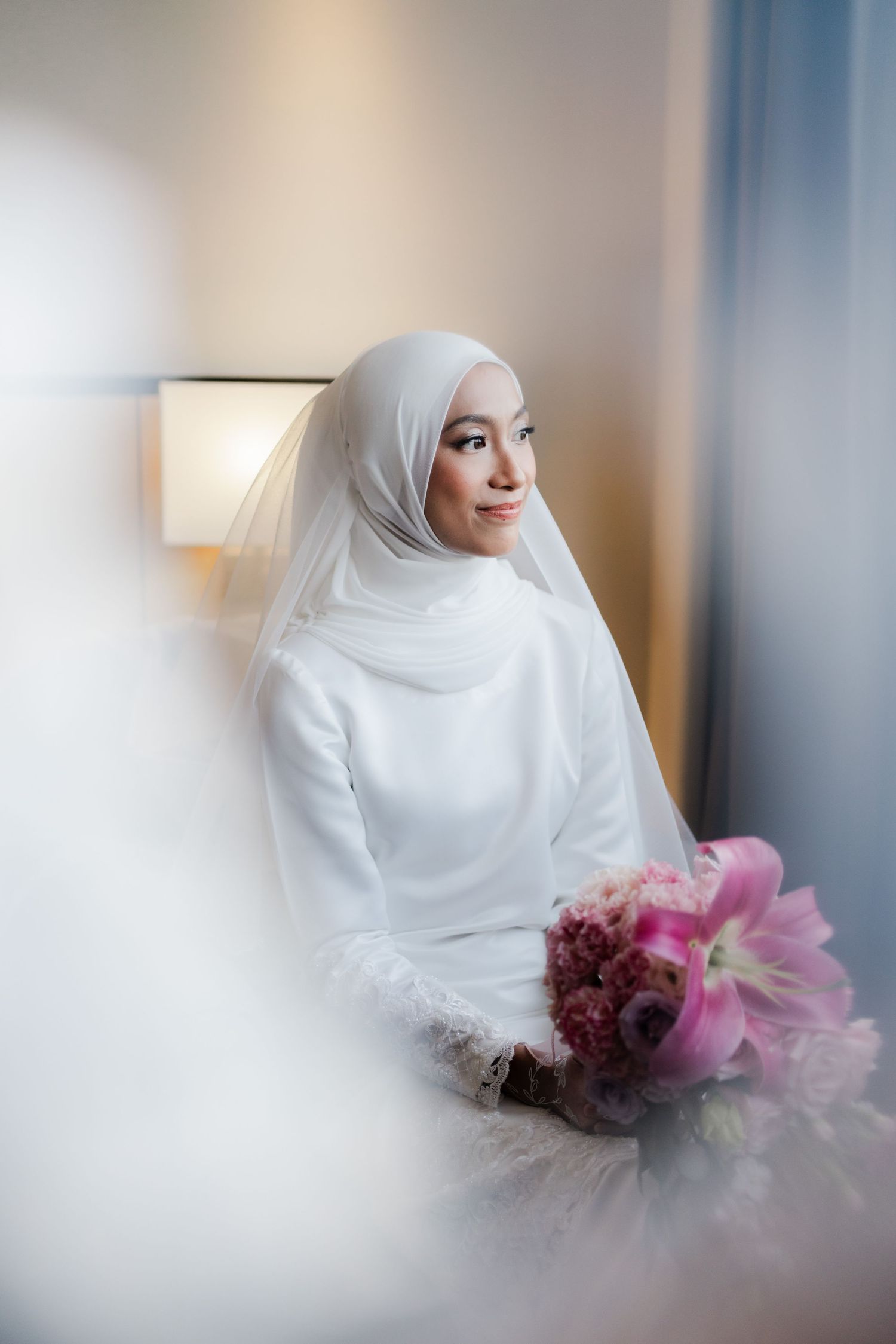 Elegant bridal portrait with soft lighting highlighting white hijab and pink floral bouquet.