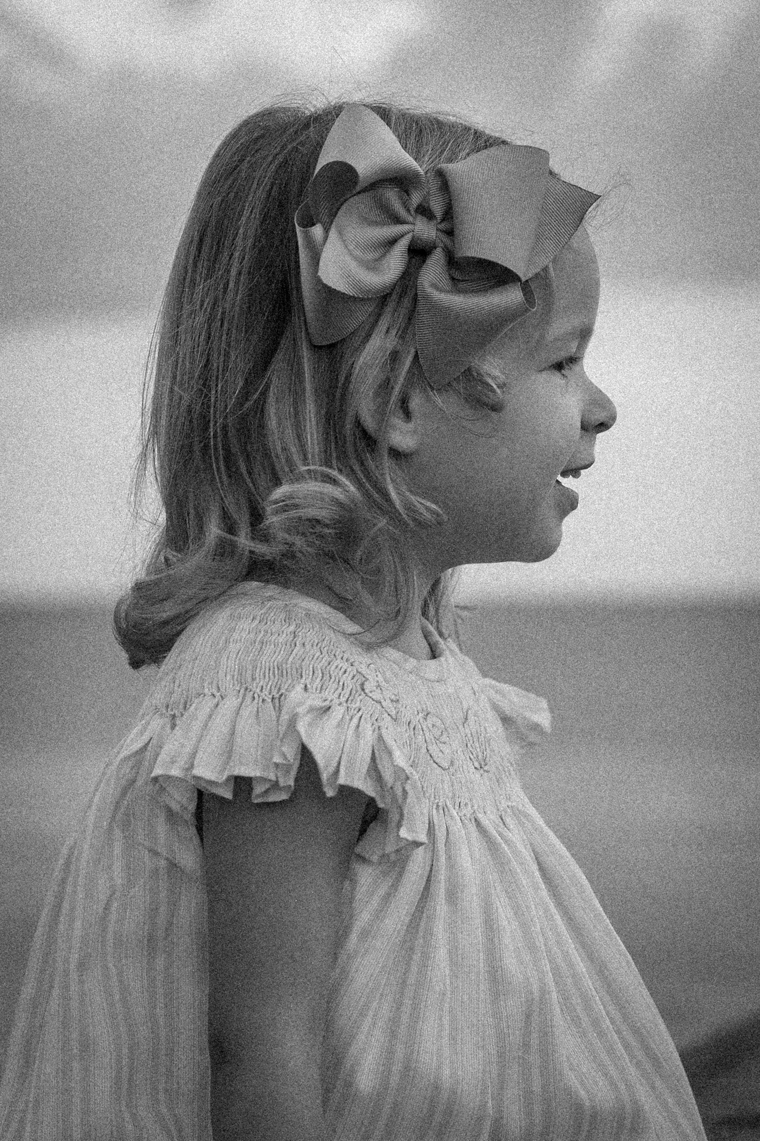 A black and white profile portrait of a young girl with a large bow in her hair near the ocean.
