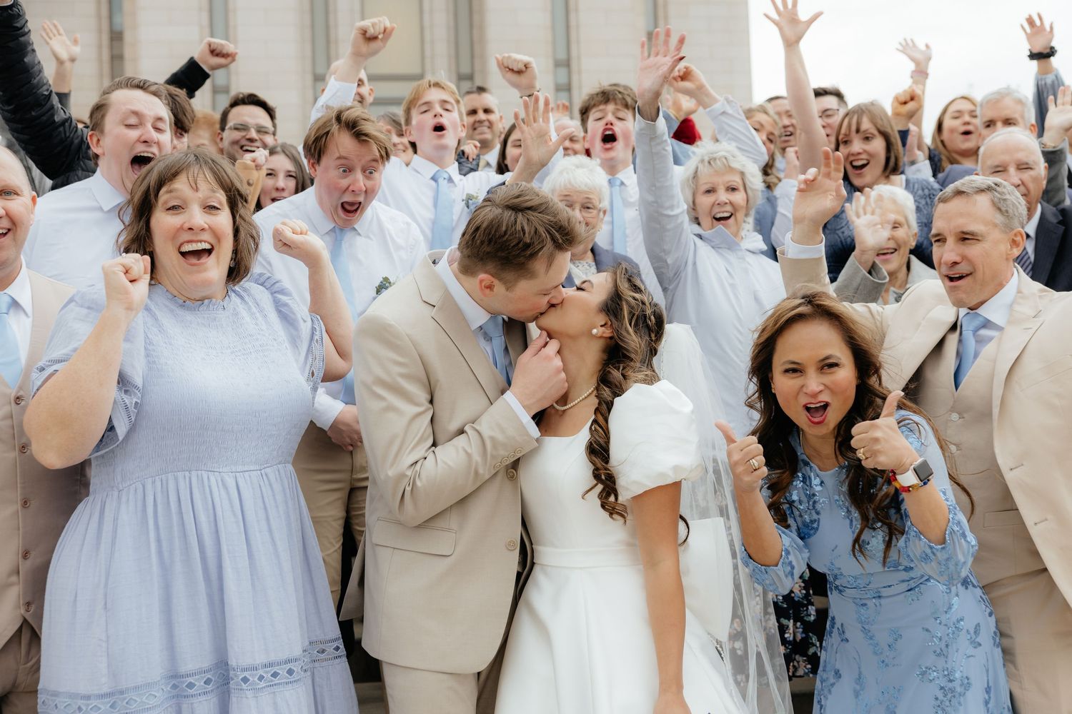 Excited wedding party and family celebrating with cheers as bride and groom kiss on steps.