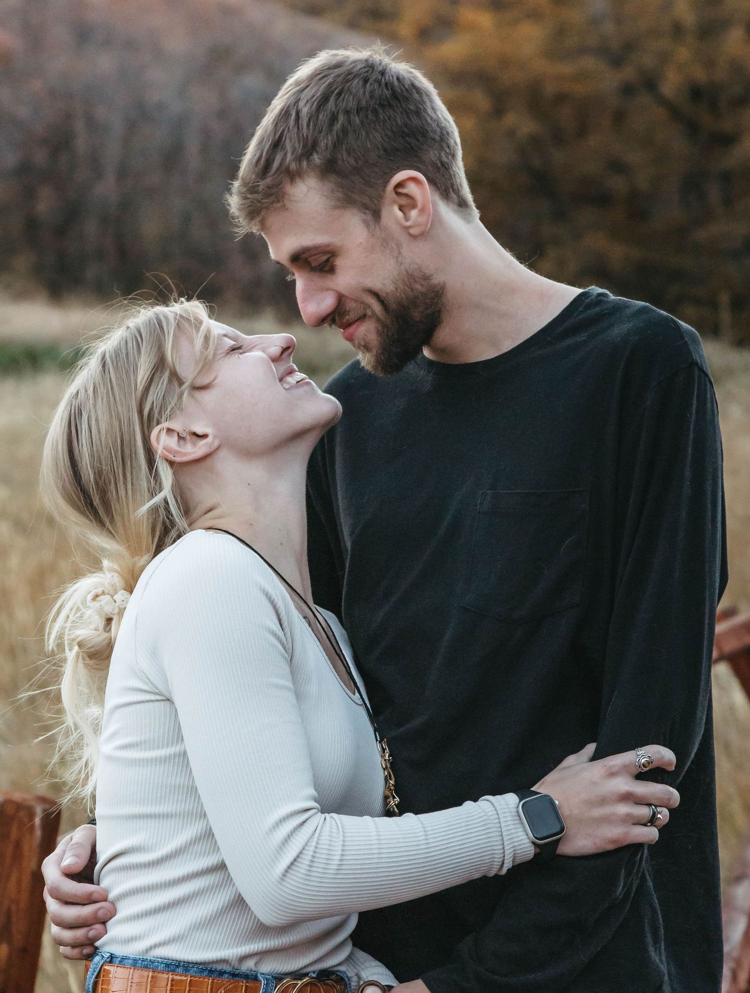 A couple shares a tender moment laughing together outdoors during autumn with warm lighting and fall colors in background.