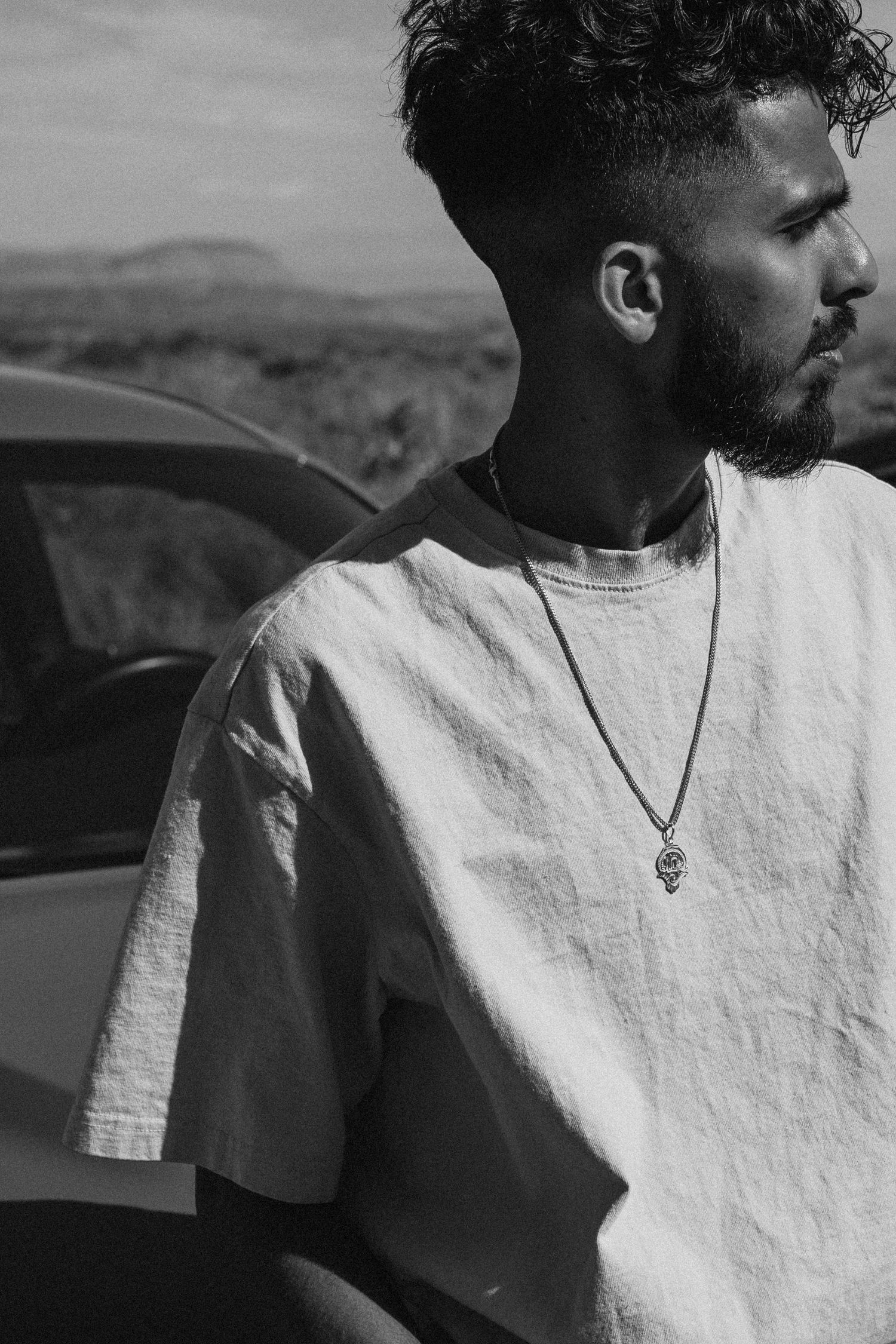 Black and white portrait of person in white t-shirt and necklace looking to the side against blurred outdoor background.