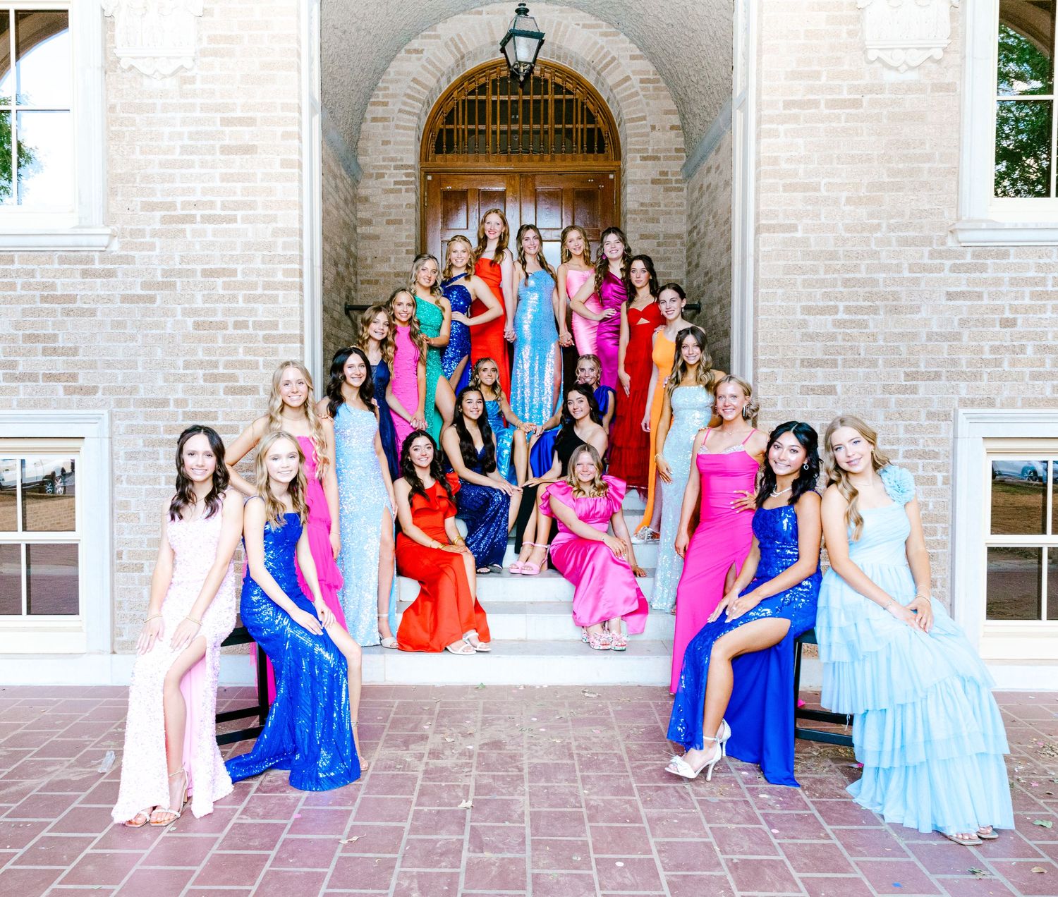 Group of women in formal gowns posing on brick steps in front of an arched doorway.