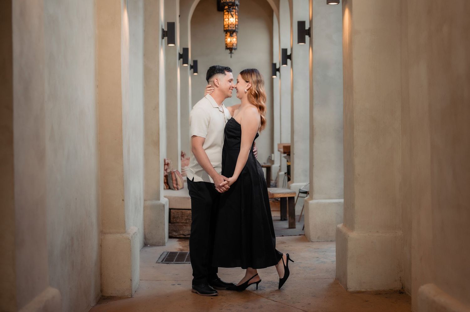 A romantic couple embraces in an elegant corridor with stone columns and hanging lanterns creating a dreamy ambiance.