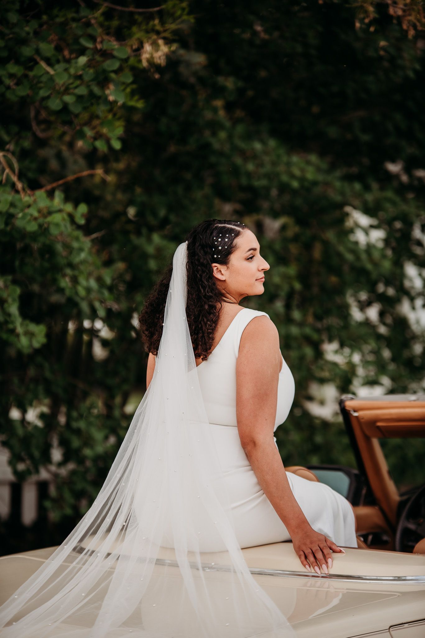 Bride in white dress and veil sitting in vintage convertible car during wedding photoshoot.