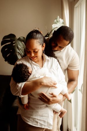 A tender family moment by a window as parents embrace their newborn baby in warm natural light.
