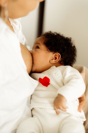 Baby in white clothing breastfeeding while being held by someone wearing white clothing.