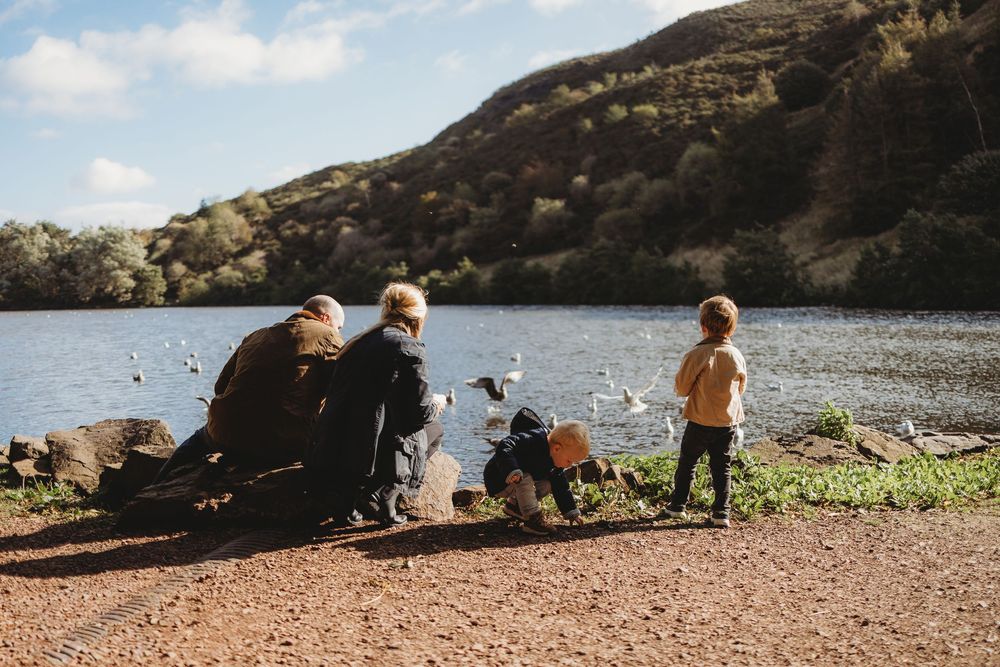 Family enjoying a peaceful afternoon by a scenic lake with mountains, watching birds on a sunny day.