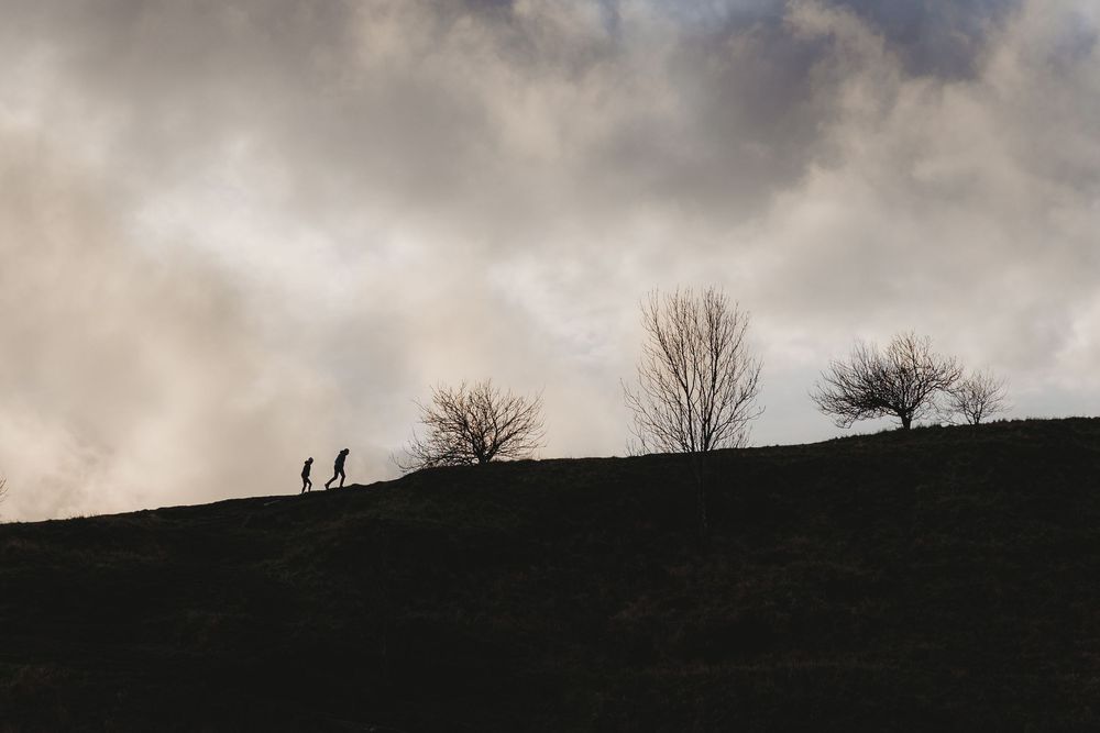 Two silhouetted figures walk along a dark hilltop ridge against dramatic cloudy skies at dusk.