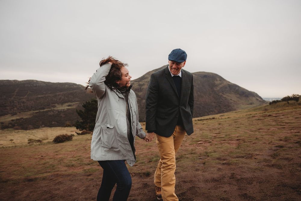 Two people share a joyful moment while hiking on a windswept hill under an overcast sky.