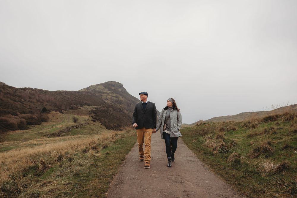 Two people walk along a scenic mountain trail with rolling hills and rugged terrain in the background.