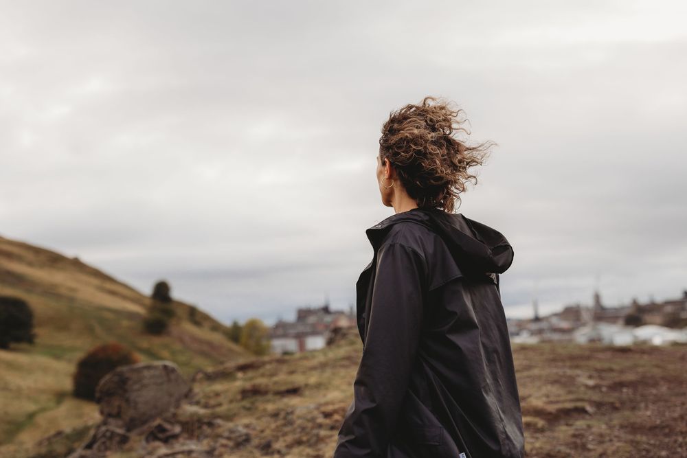 Person with windswept hair sits on rocky terrain overlooking a misty coastal landscape at sunset.