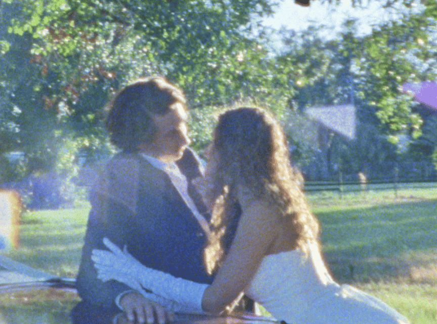 A couple in wedding attire dances together in a sunlit outdoor garden setting.