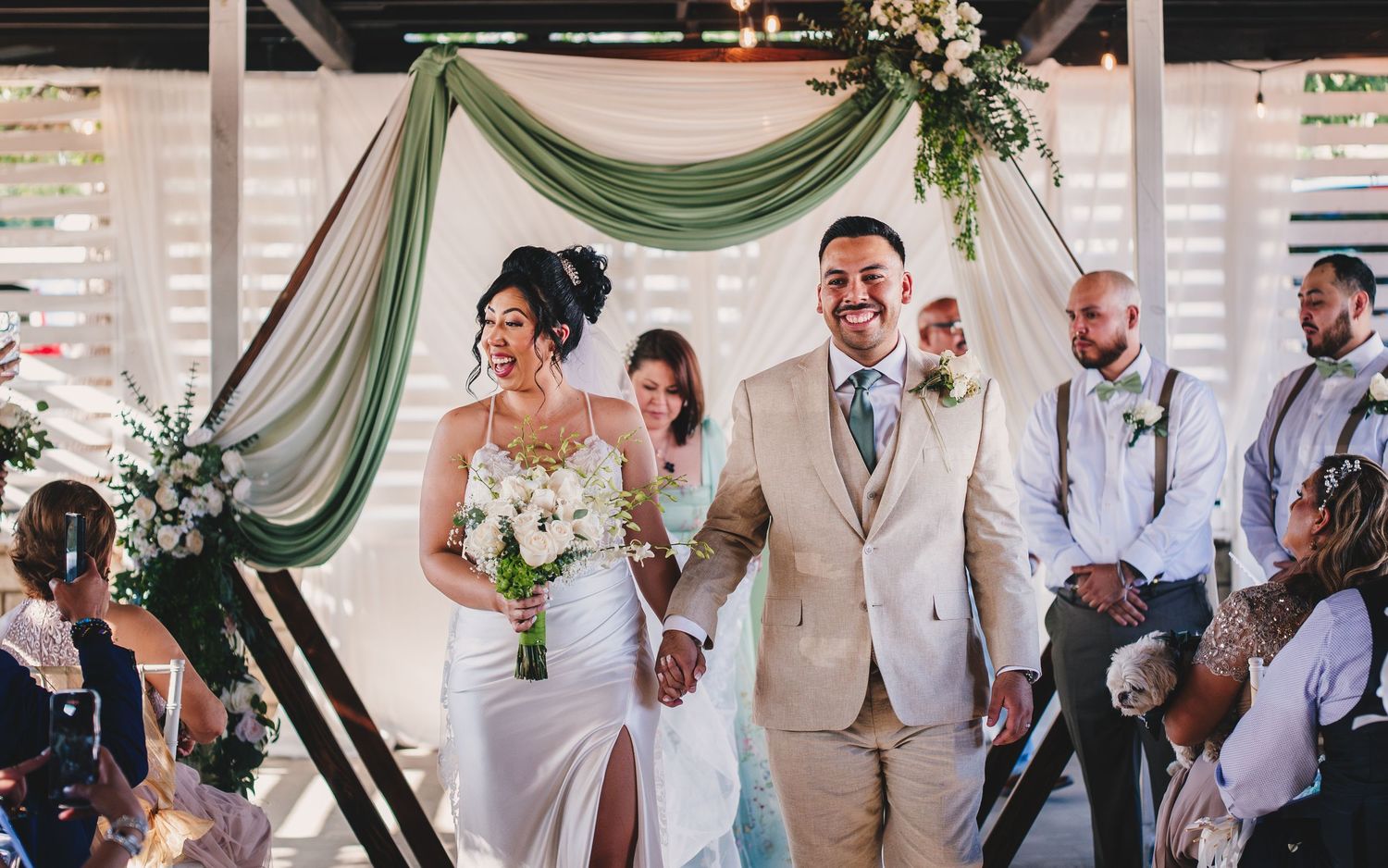 Wedding party celebrates with champagne toasts under elegant green and white draped ceremony backdrop.