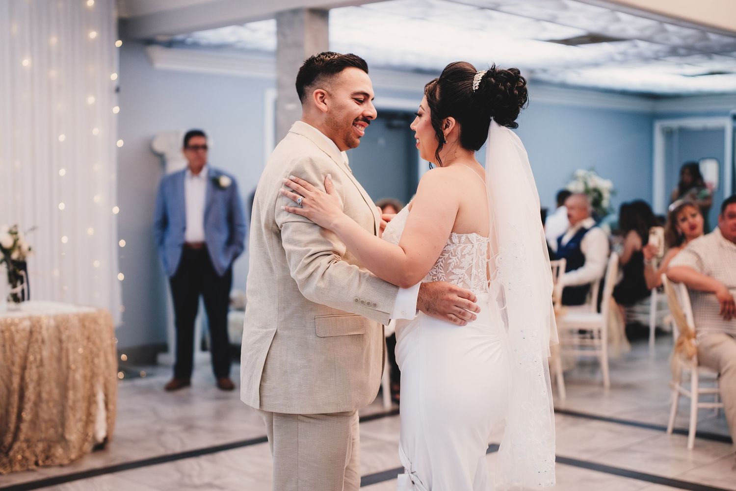 A couple shares their first dance at a wedding reception in a room decorated with twinkling lights and gold accents.