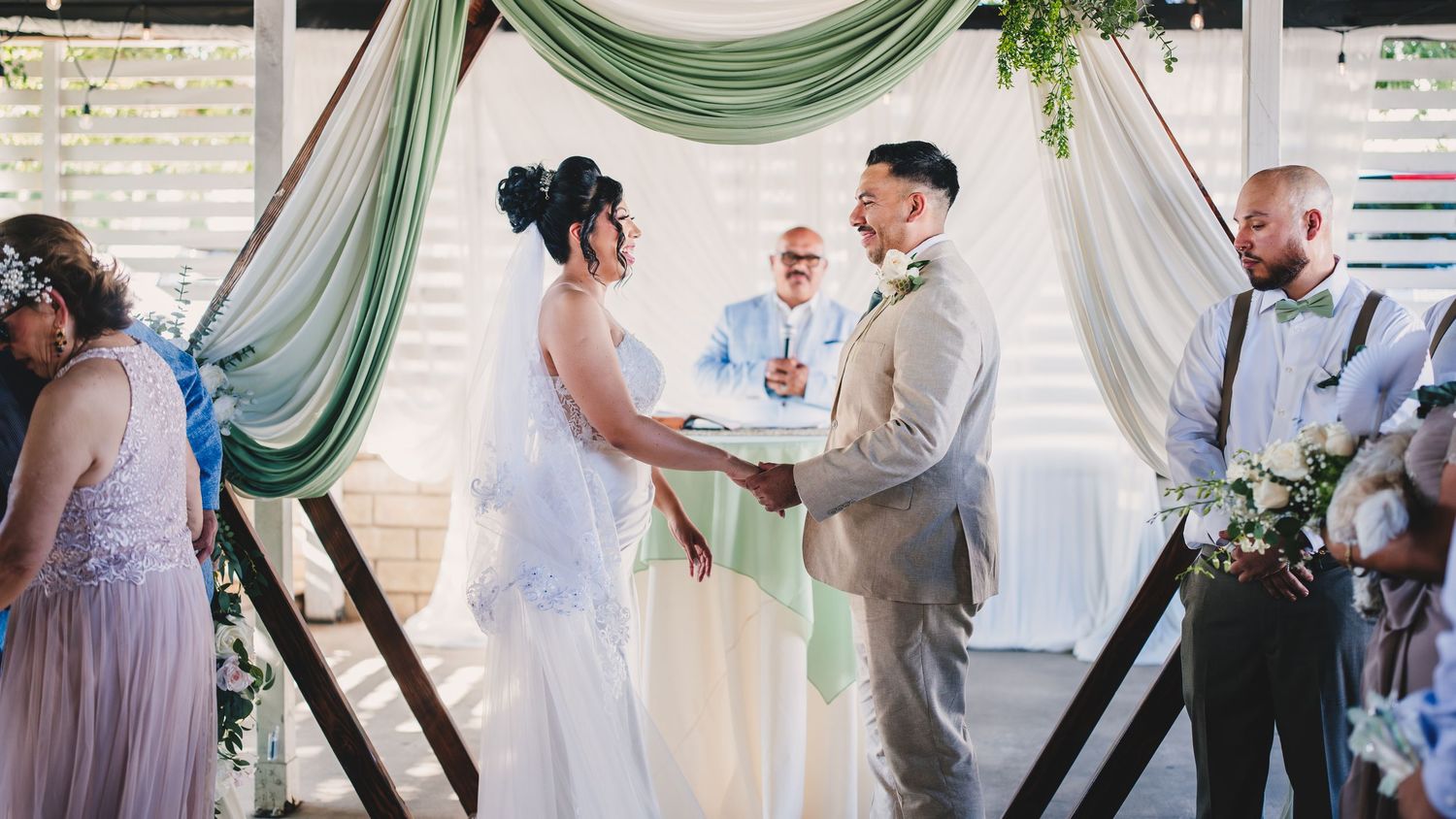 Wedding party gathers in an elegant reception venue decorated with soft green drapery and natural light.