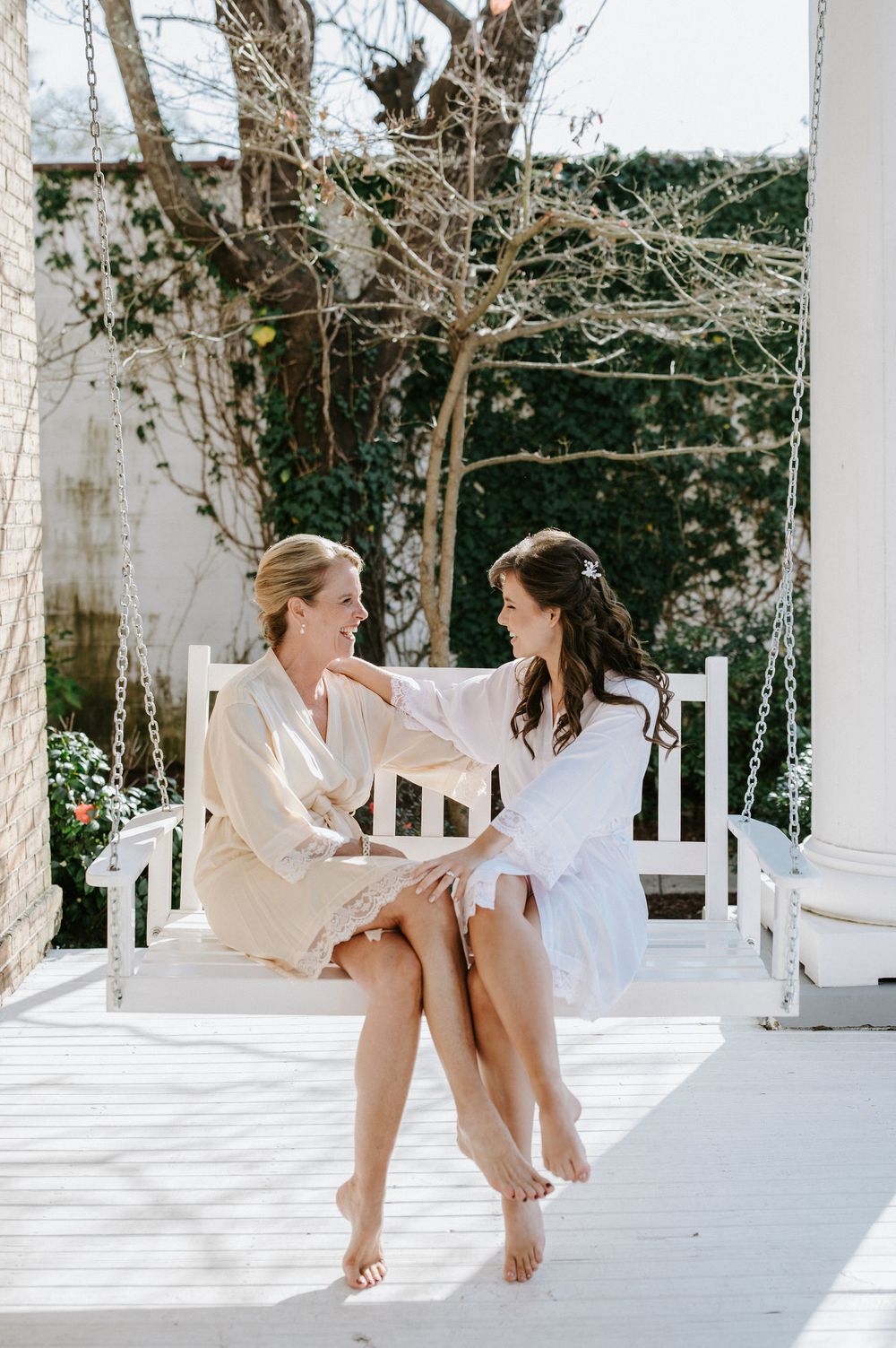 Two women in white robes sit together on a white porch swing in a garden setting with bare trees.
