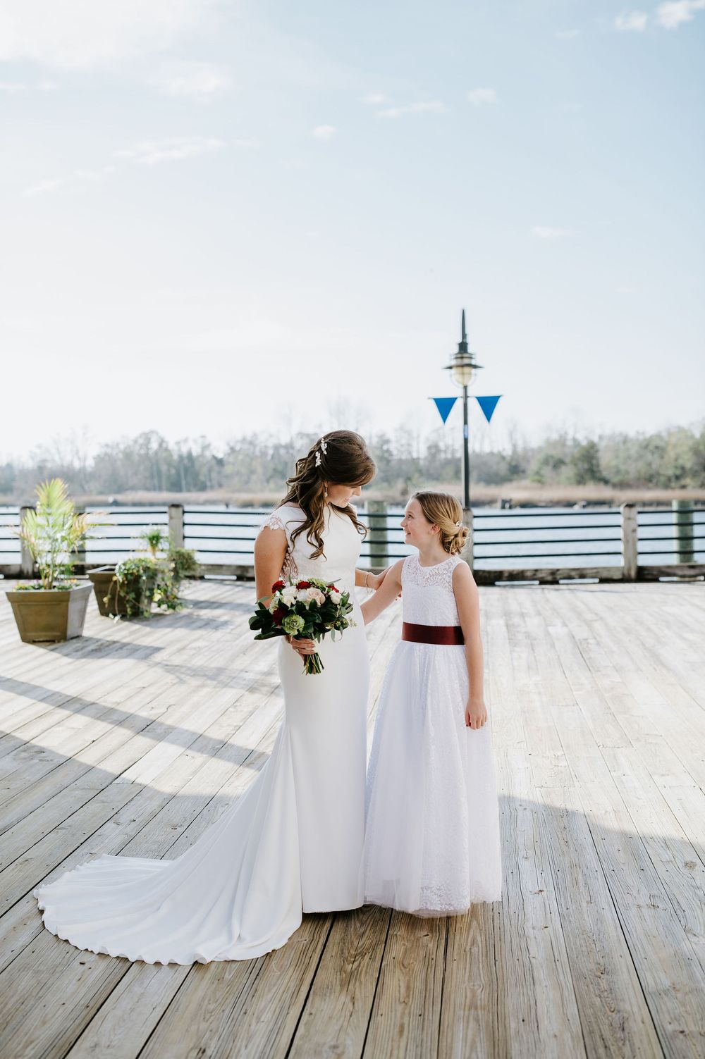 Two people in white wedding dresses sit together on a wooden deck overlooking a scenic waterfront view.