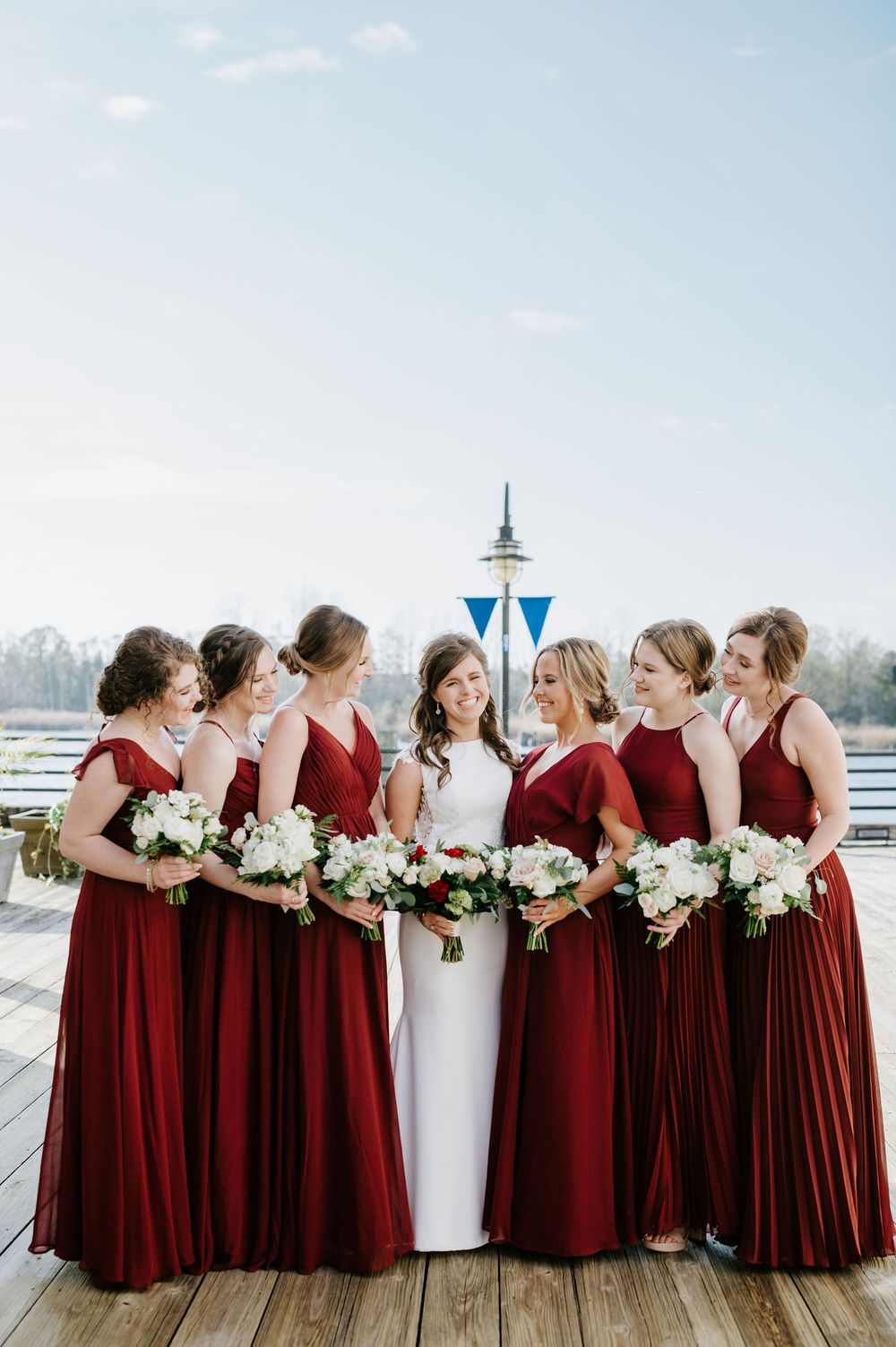 Bridal party in burgundy red bridesmaid dresses and white wedding gown pose together on wooden deck overlooking marina.