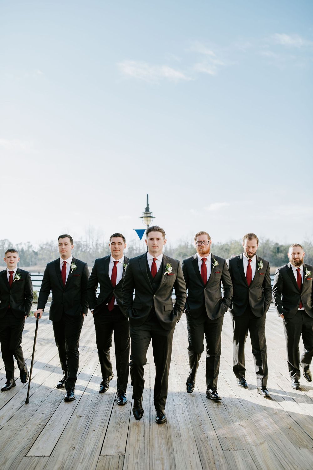 Groomsmen in black suits and red ties pose playfully on a wooden deck during a winter wedding photo shoot.
