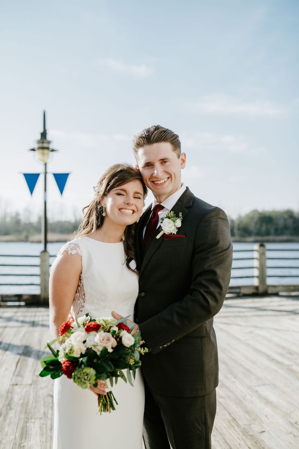 A couple in wedding attire stands on a wooden pier with a bouquet of red and white flowers on a sunny day.