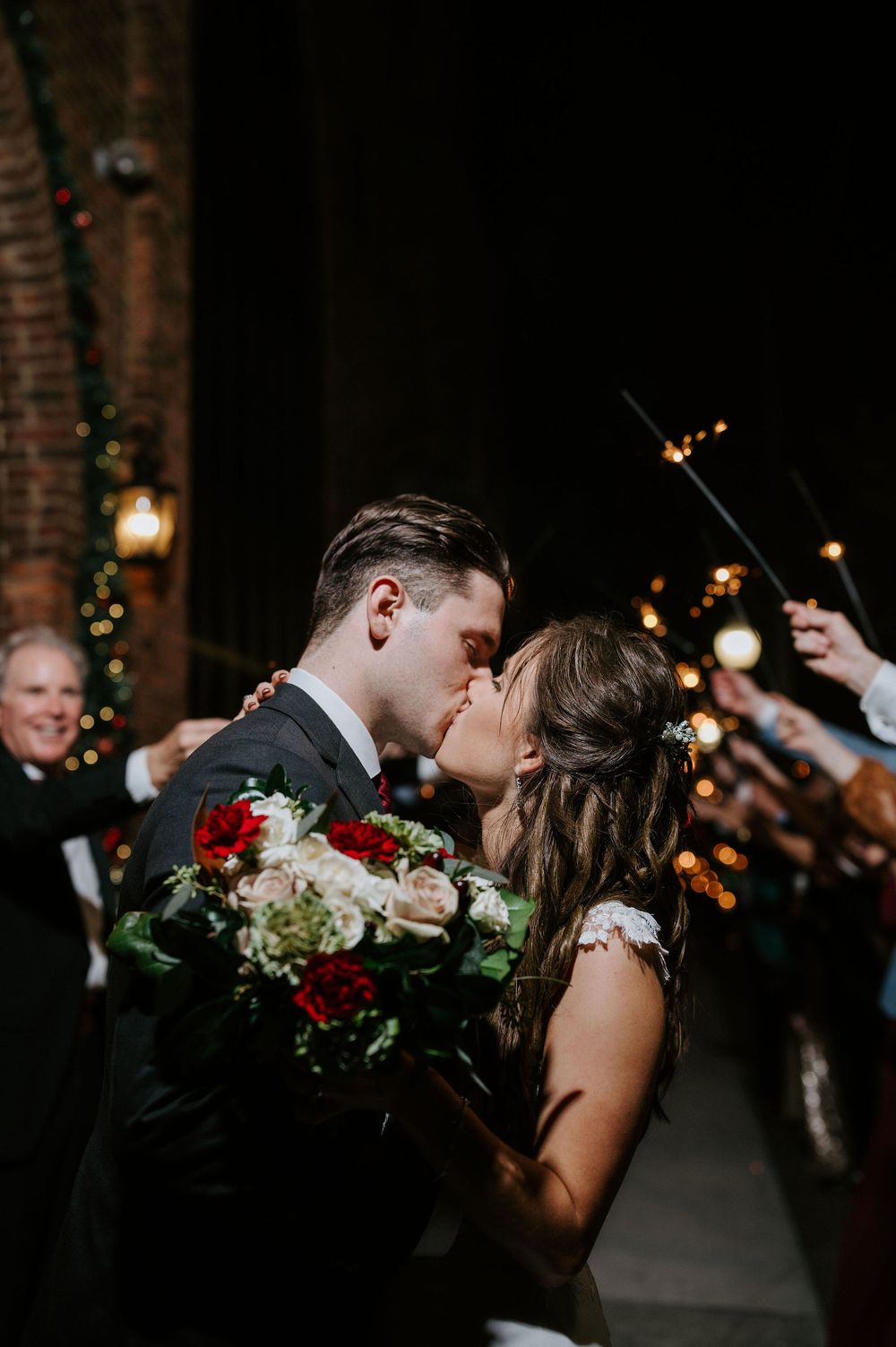 A romantic wedding moment with sparklers glowing in the dark as the couple shares a kiss while holding red and white flowers.