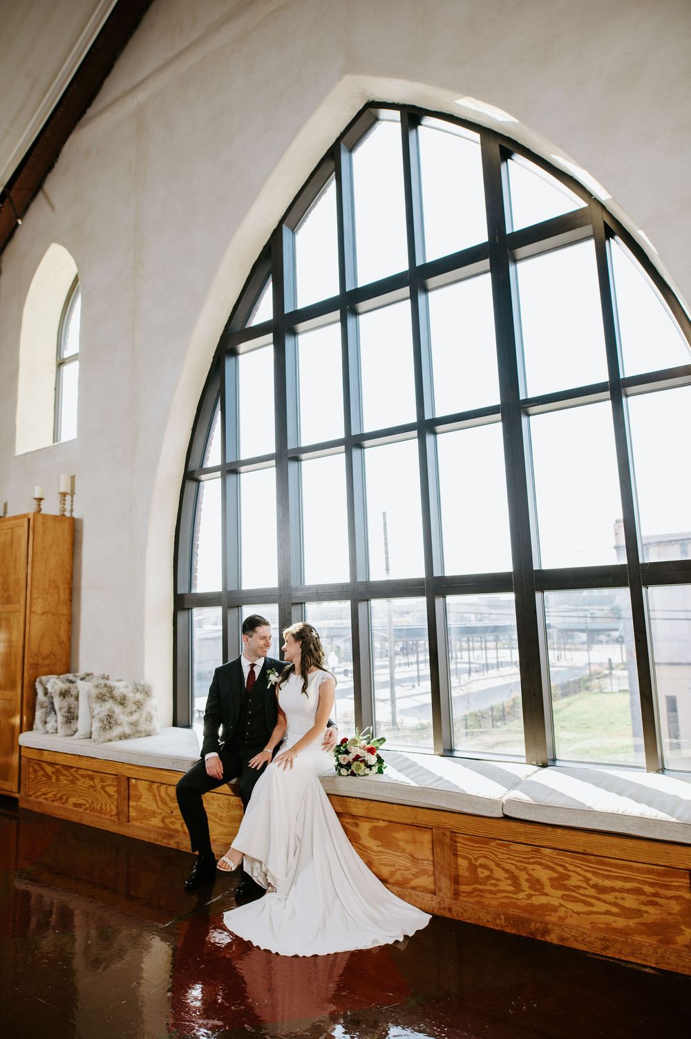 A wedding couple poses by large arched windows in a bright venue with wooden benches and white walls.