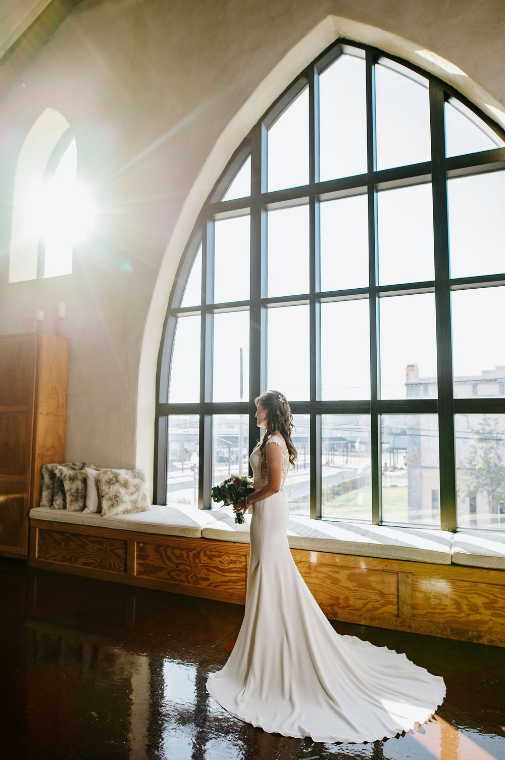 Elegant white wedding gown silhouetted against a dramatic arched window in a church venue with wooden details.