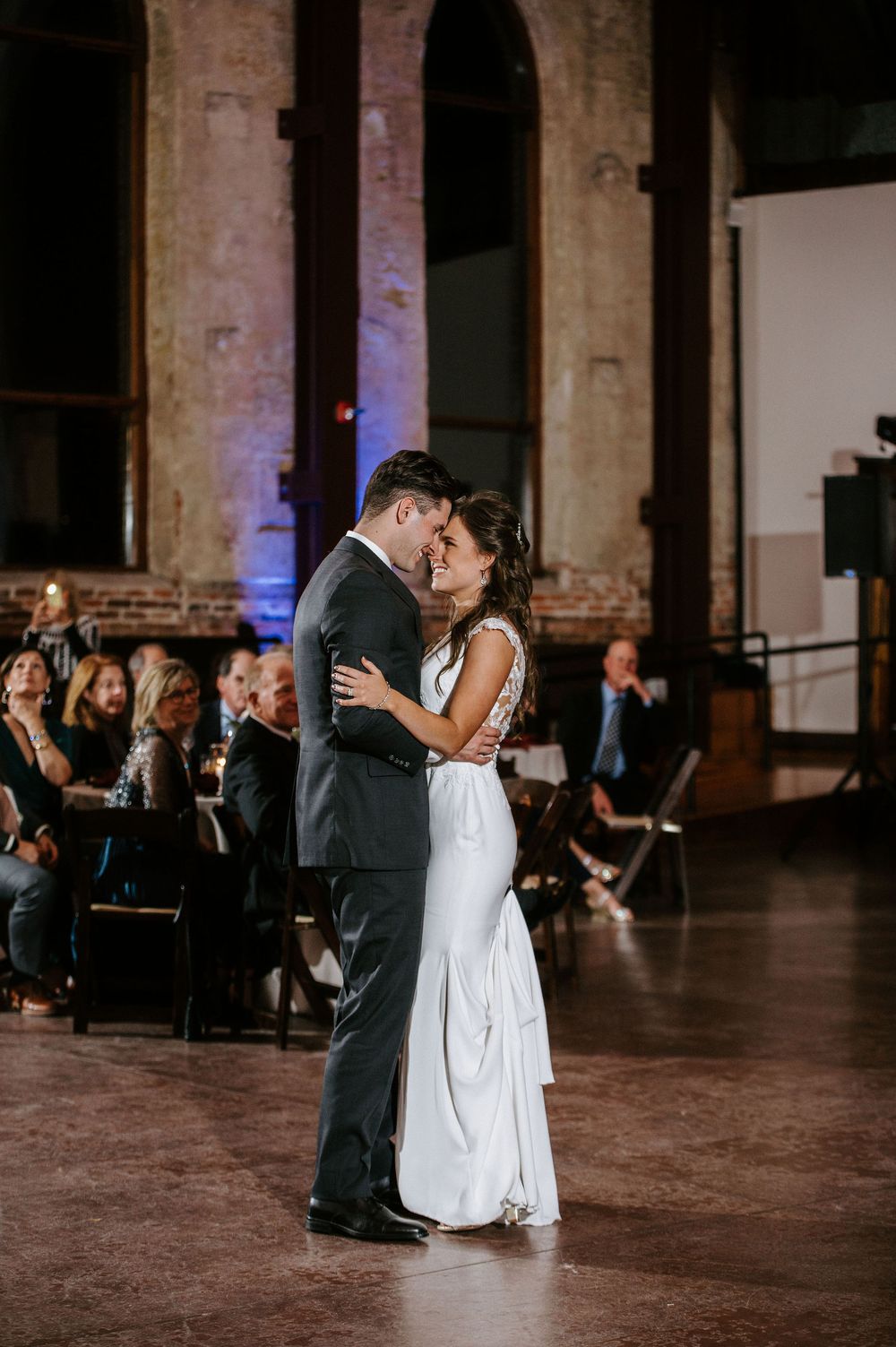 A couple shares their first dance in an elegant historic venue with exposed brick walls and ambient lighting.