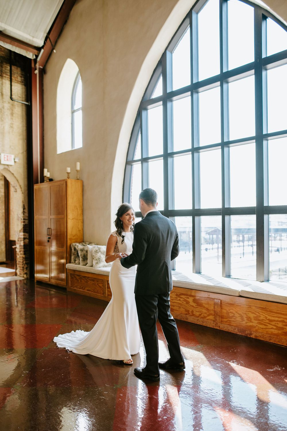 A couple in wedding attire shares a tender moment in a sunlit room with arched windows and hardwood floors.